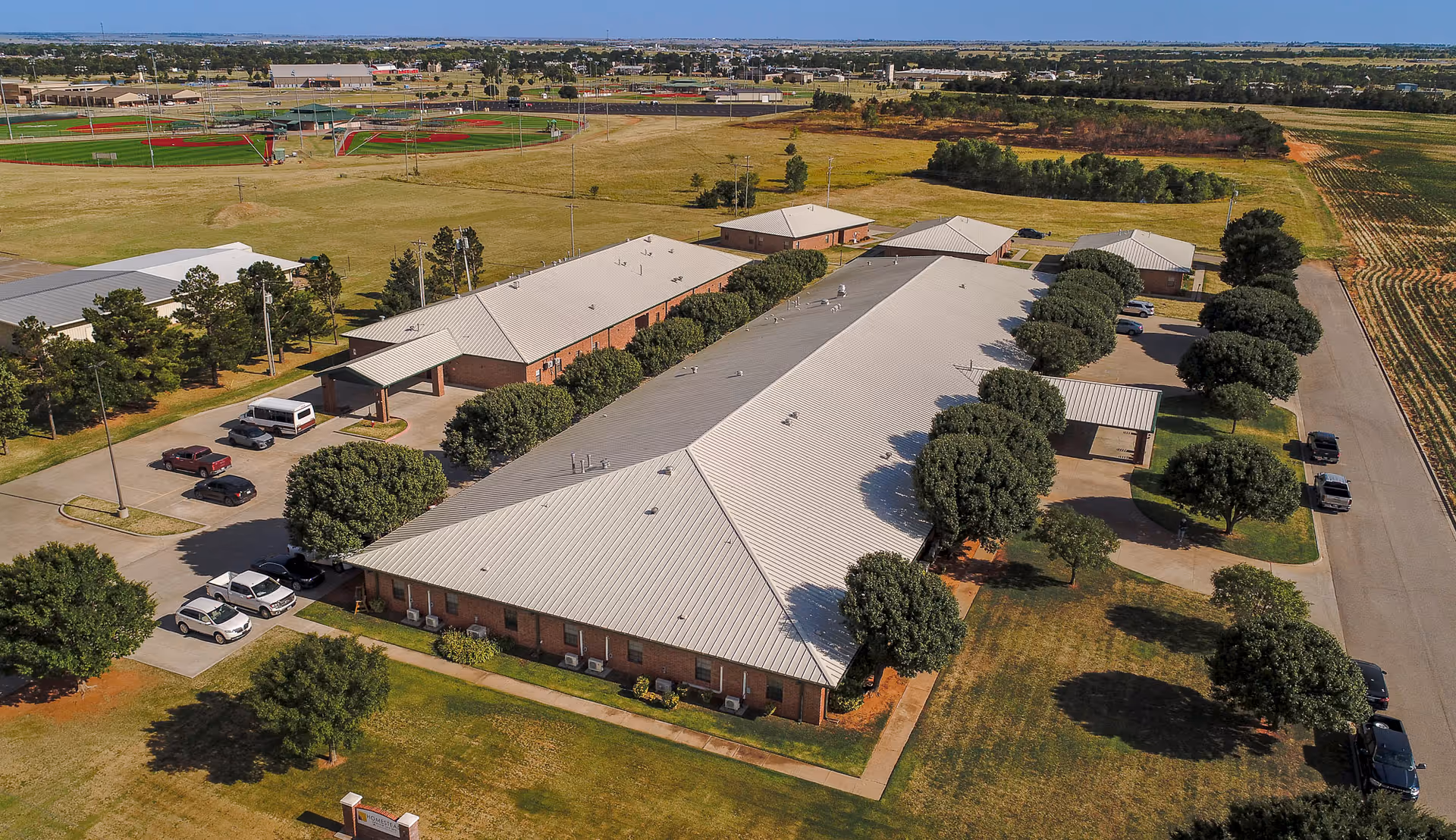 Aerial view of a single-story senior living facility with a light-colored metal roof, surrounded by trees and parking areas. The building is situated in a rural area with open fields and sports fields visible in the background.