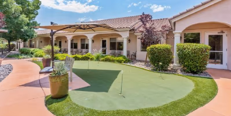 Outdoor courtyard area of a senior living facility with a small putting green, patio umbrella, chairs, potted plants, and a building with a tiled roof and arched windows in the background under a blue sky with some clouds.