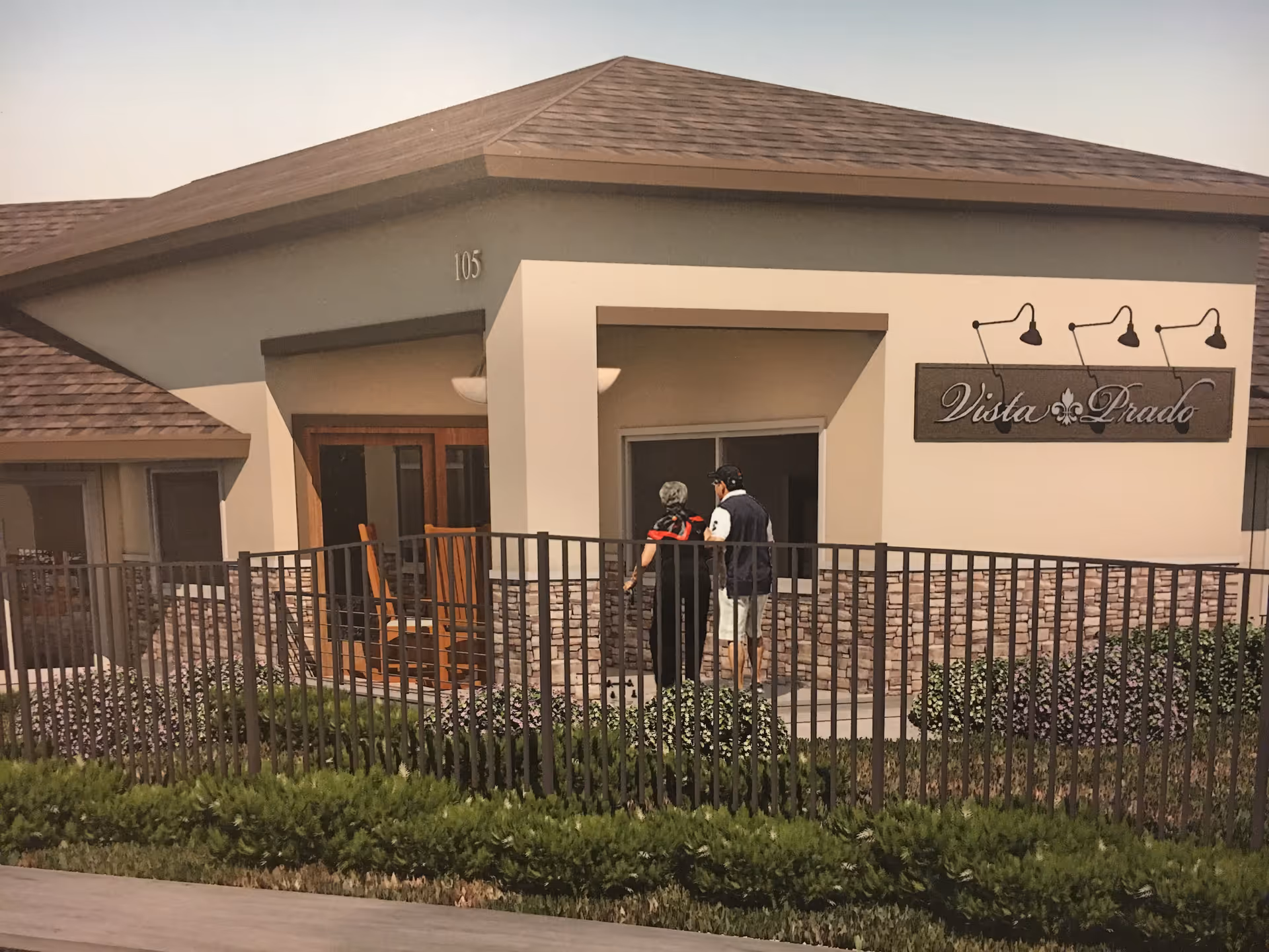 Front entrance of the Vista Prado building showing a fenced patio, two people standing by the doorway, and a mounted Vista Prado sign.