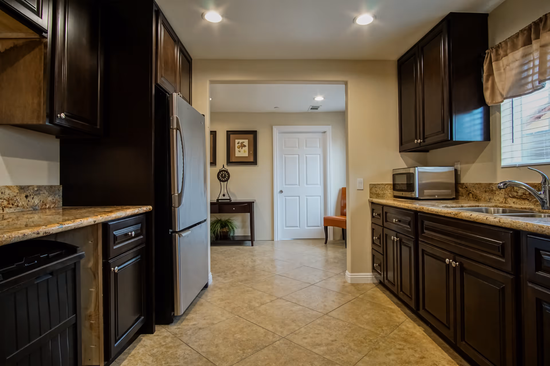 Interior view of a kitchen with dark wooden cabinets, granite countertops, a stainless steel refrigerator, a microwave, and a double sink. The floor is tiled, and there is a doorway leading to another room with a white door, a small table with a plant, framed artwork on the wall, and an orange chair.