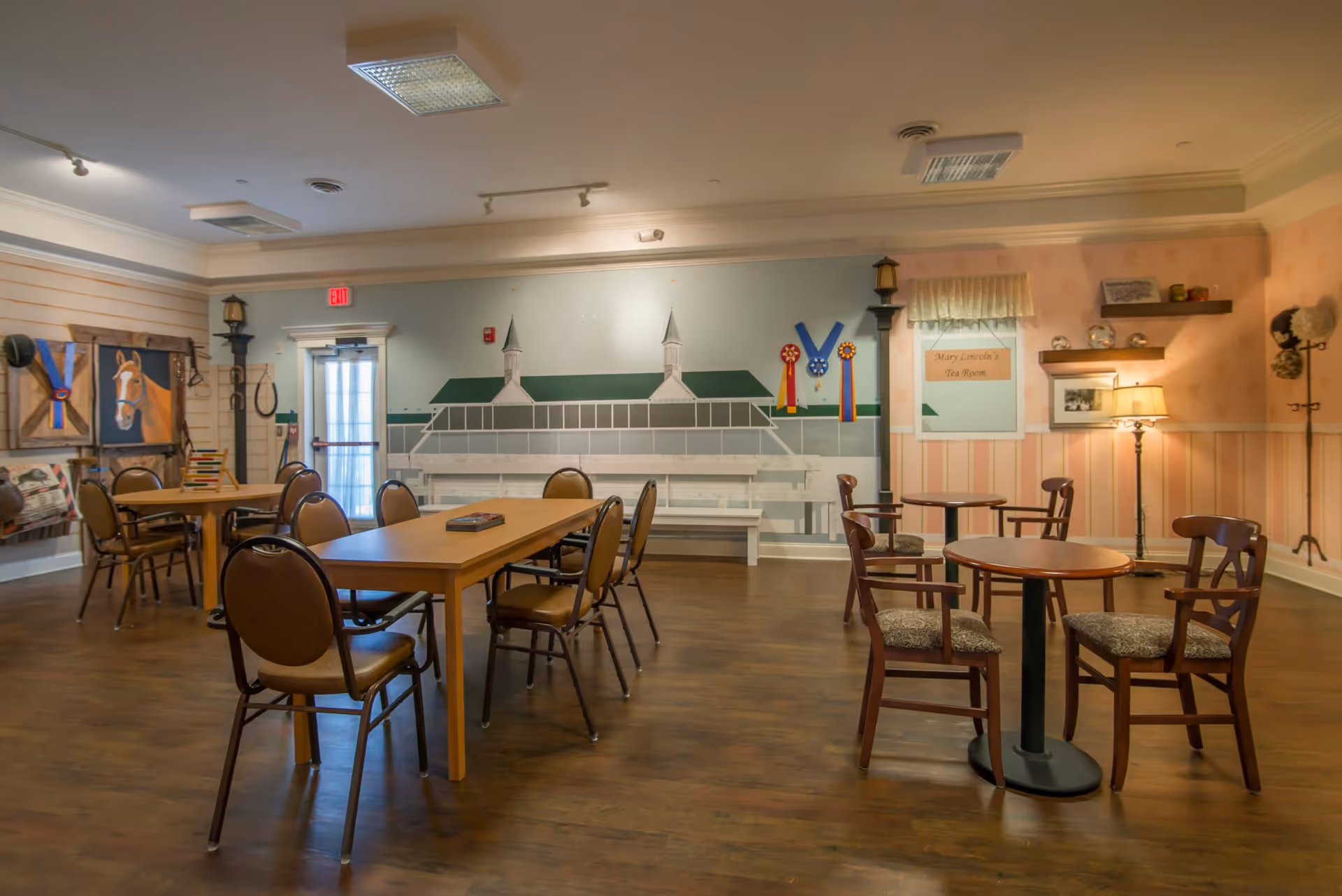 A cozy room with wooden floors and several tables and chairs arranged for seating. The walls are decorated with horse-themed artwork, ribbons, and a mural of a building with two cupolas. A sign on the wall reads 'Mary Lincoln's Tea Room'. The room is softly lit with ceiling lights and a floor lamp, creating a warm and inviting atmosphere.