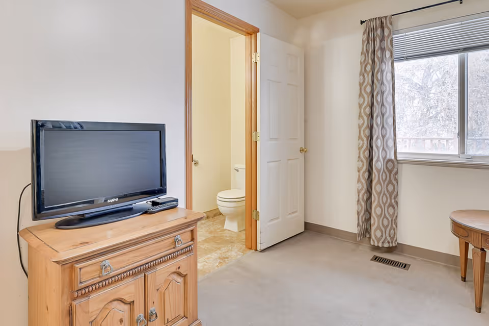 A room with a wooden cabinet holding a flat-screen TV and remote, an open door leading to a bathroom with a visible toilet, a window with patterned curtains, and a small wooden table in the corner.