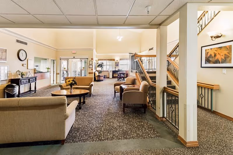 A spacious and well-lit common area in a senior living facility with beige sofas and armchairs arranged around a wooden coffee table with a flower vase. The room features carpeted flooring, a staircase with wooden handrails and decorative iron balusters, and framed artwork on the walls. In the background, there is a reception desk, entrance doors, and additional seating areas near windows.