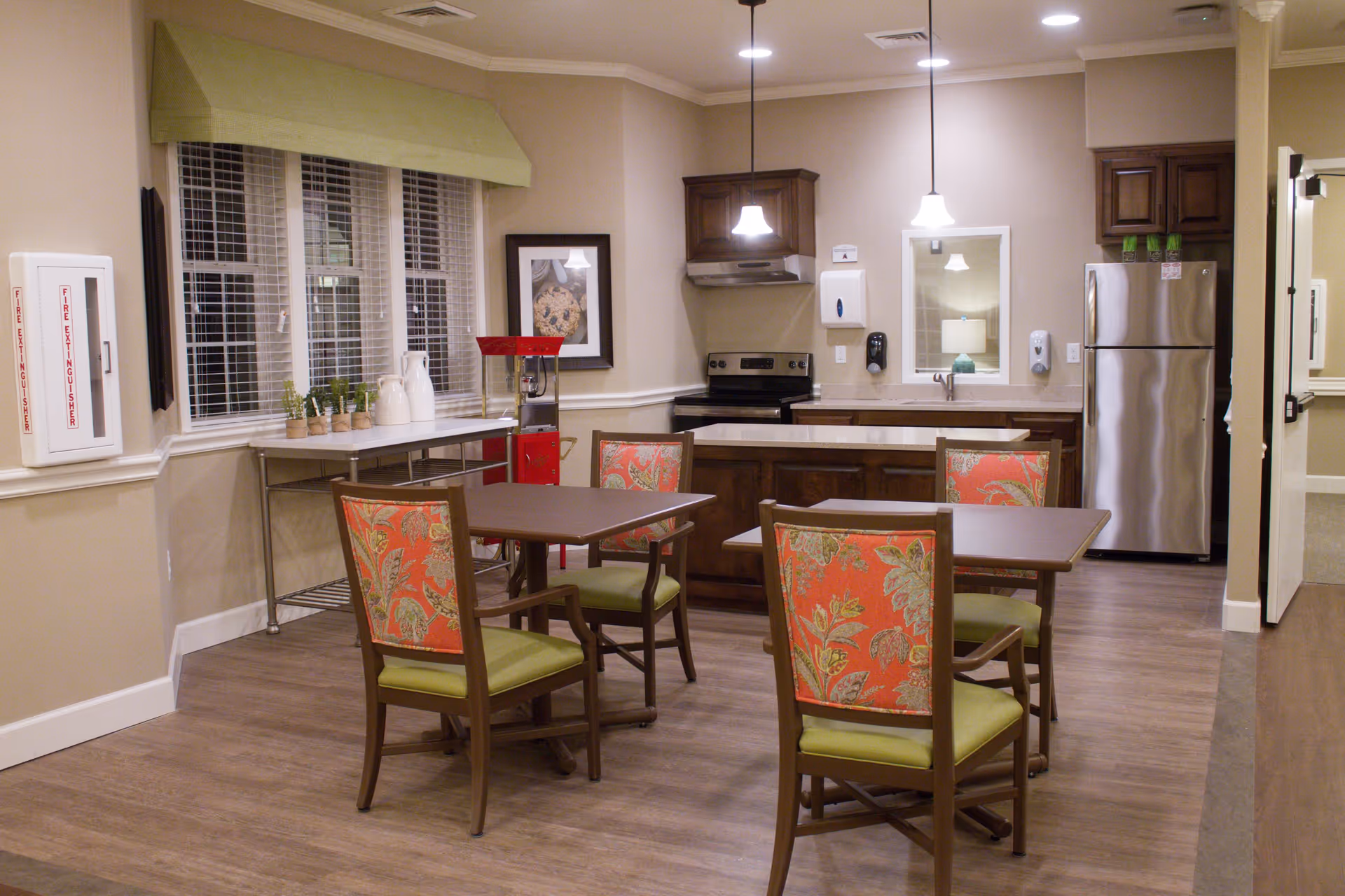 A cozy kitchen and dining area in a senior living facility with wooden cabinets, stainless steel refrigerator and stove, a kitchen island, and two tables with chairs featuring floral patterned upholstery. The room has wood flooring, beige walls, and a window with blinds and a green valance.