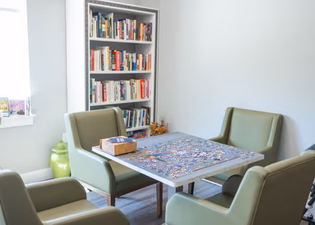 A small room with a square table in the center covered with a partially completed jigsaw puzzle. Four green cushioned chairs surround the table. In the background, there is a white bookshelf filled with books. A window on the left side lets in natural light, and a green decorative vase is placed near the window.
