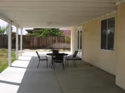 Covered outdoor patio with a round table and four chairs on a concrete slab beside a house and fenced yard.