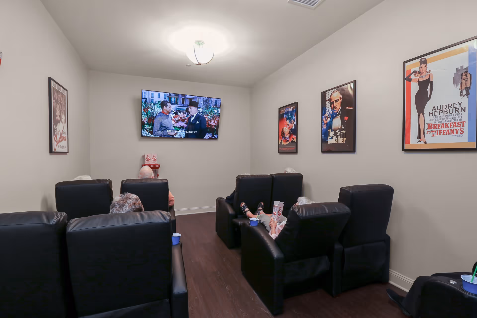 A small movie theater room with six black leather recliners arranged in two rows facing a wall-mounted flat screen TV. Several elderly people are seated, watching a movie with subtitles. The walls are decorated with framed classic movie posters including Casablanca, Gone with the Wind, The Godfather, and Breakfast at Tiffany's. There is a popcorn machine on a small table beneath the TV.