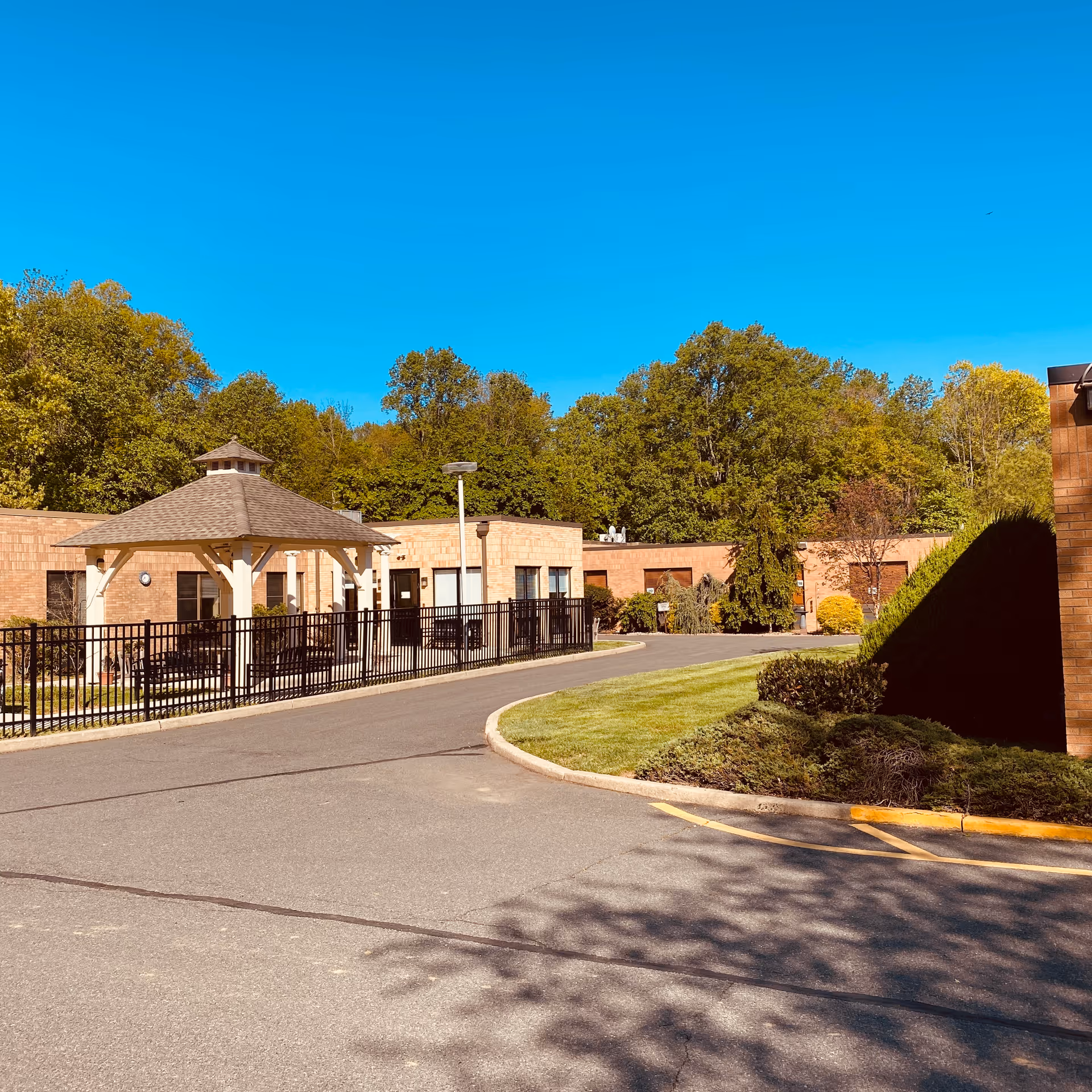 Outdoor view of Greenwood House Senior Healthcare facility showing a paved driveway, a small gazebo with a shingled roof, surrounded by a black metal fence, and a single-story brick building with windows. There are green trees and bushes in the background under a clear blue sky.