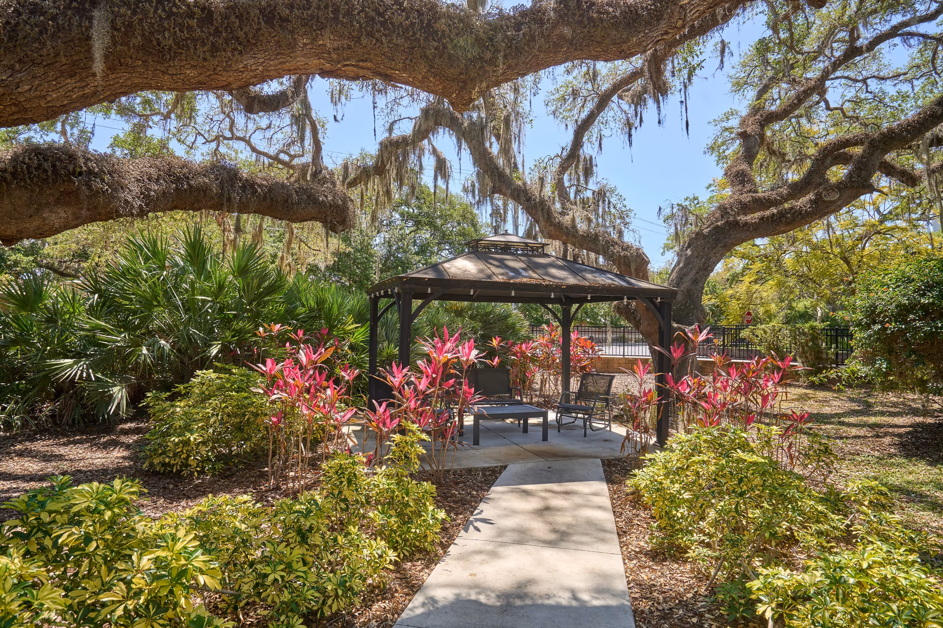 A peaceful outdoor garden area with a paved walkway leading to a gazebo surrounded by lush greenery and colorful plants. Large trees with hanging moss provide shade over the seating area inside the gazebo, which contains chairs and a table.