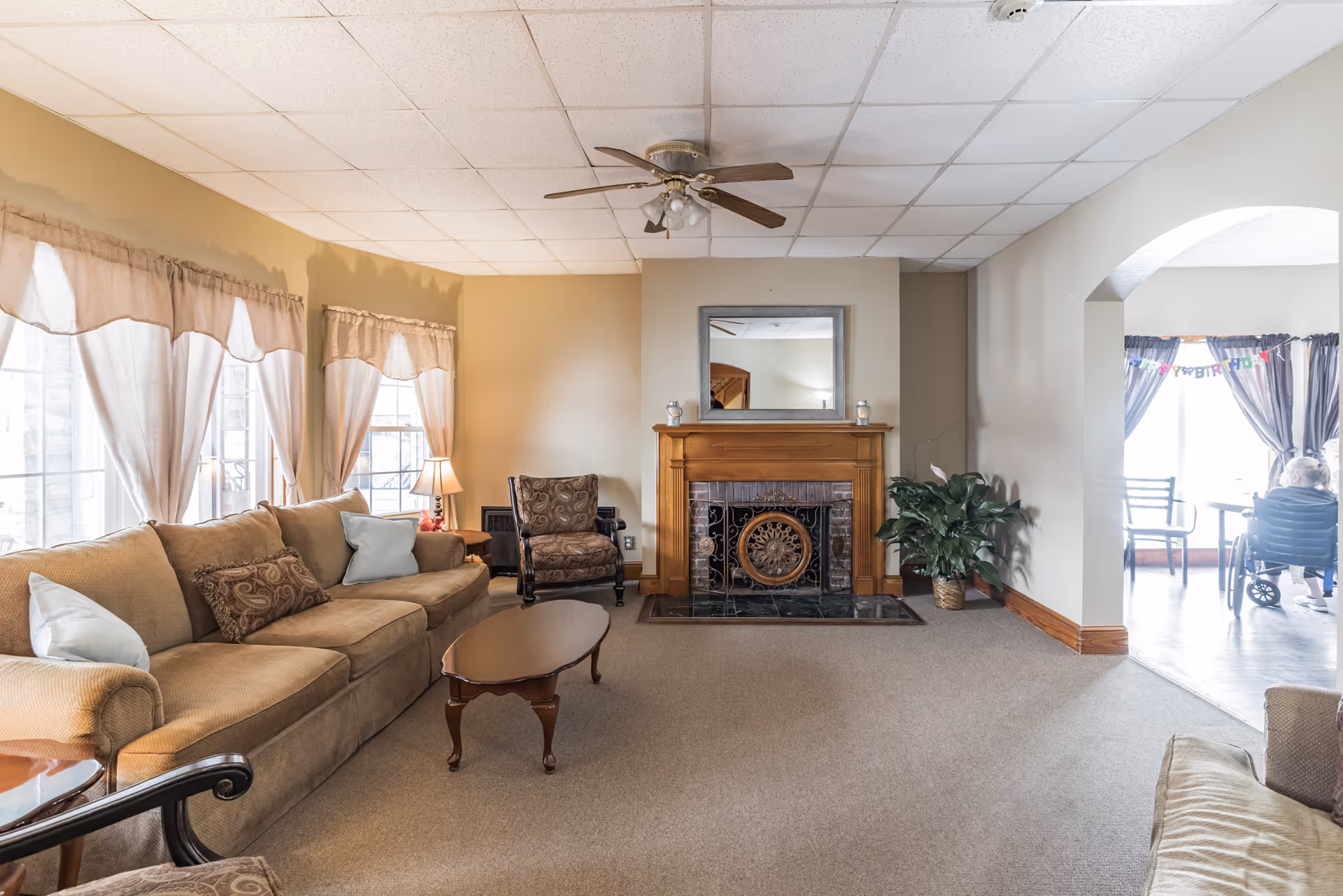 A cozy senior living facility common area with a beige sofa, armchair, wooden coffee table, and a fireplace with a mirror above it. Large windows with sheer curtains let in natural light. A ceiling fan is mounted on the white tiled ceiling. In the adjacent room, a person in a wheelchair is visible near a table with chairs and a 'Happy Birthday' banner hanging on the window.