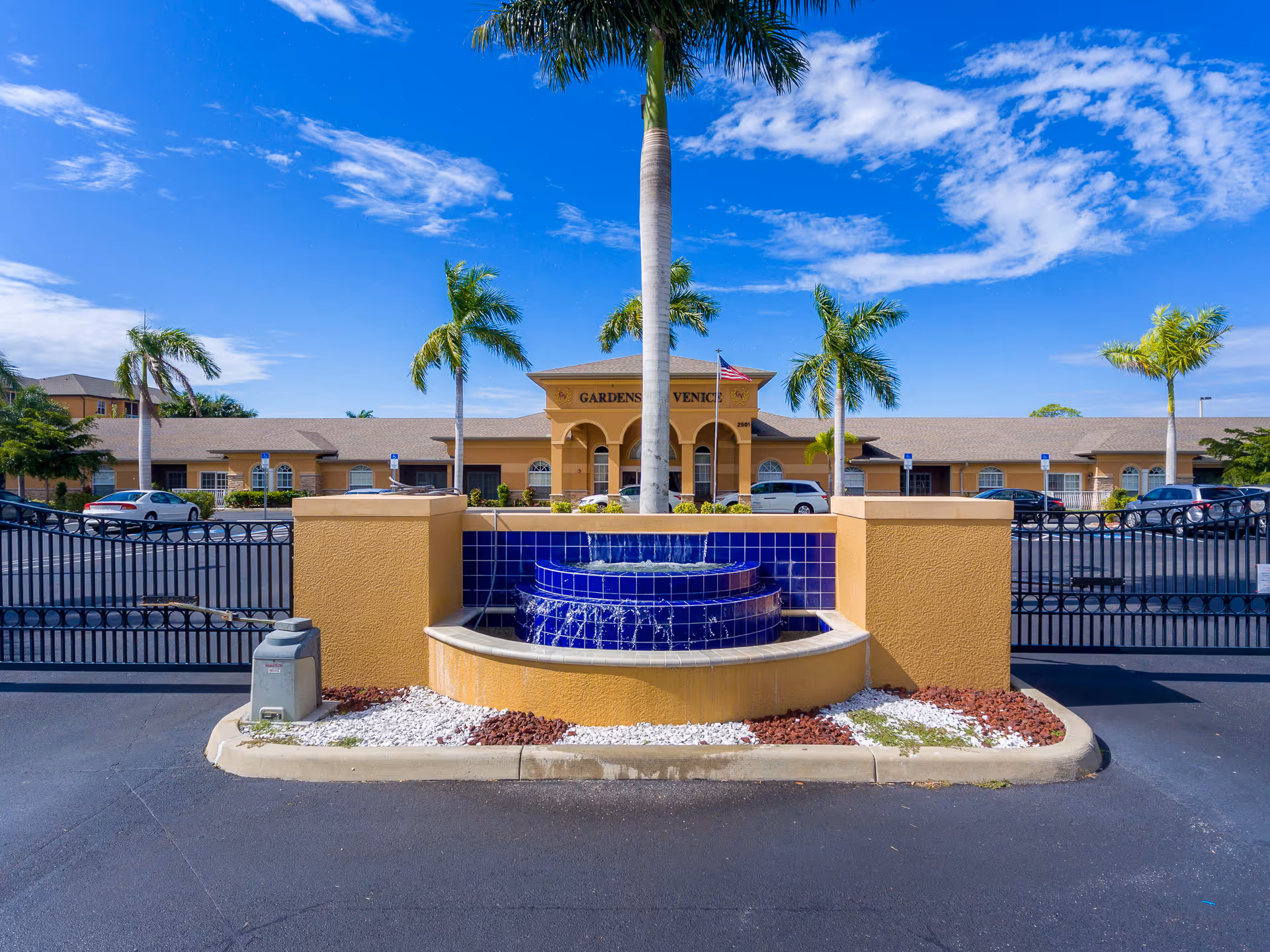 Entrance of the Gardens of Venice senior living facility with a blue-tiled fountain, gated drive, palm trees, and the building facade.