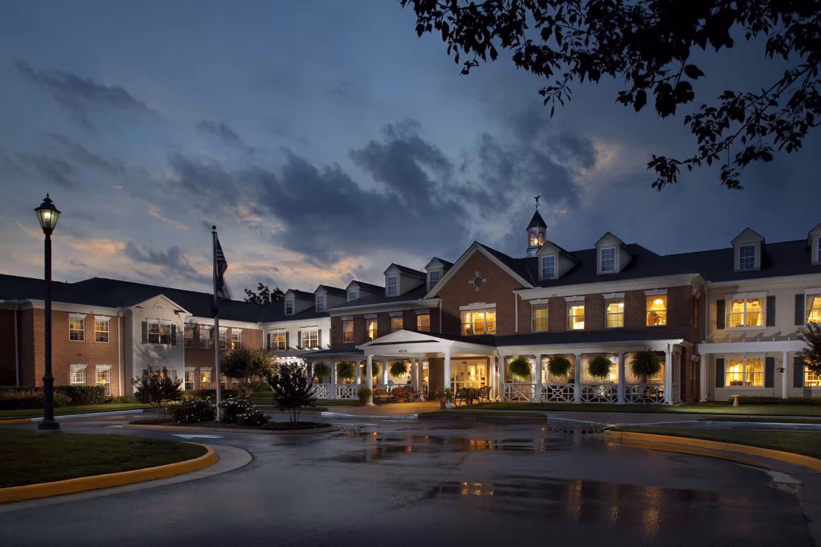 Front exterior of a large brick senior living building at dusk with lit windows, a covered entrance, flagpole, and wet driveway.