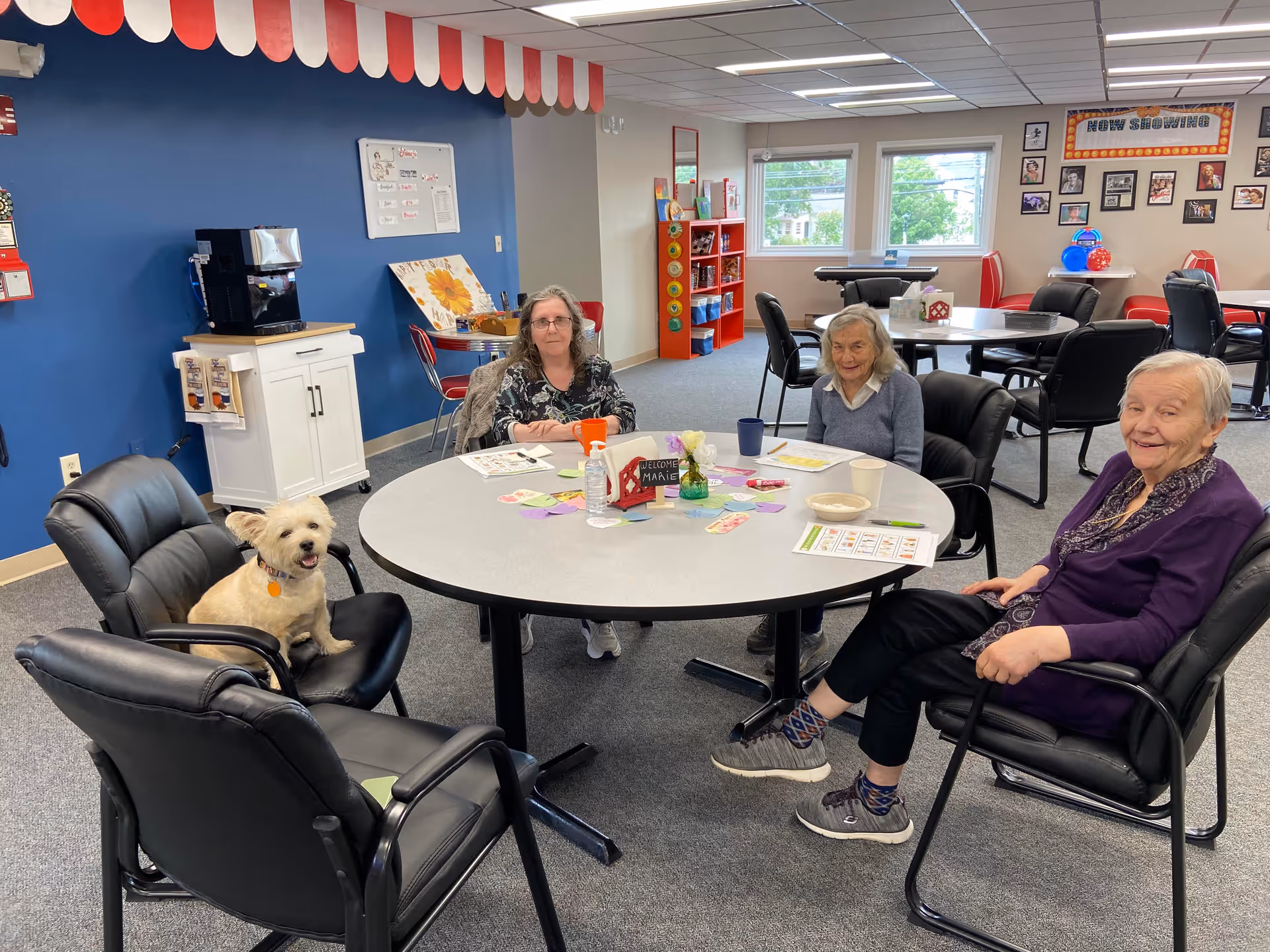 Three elderly women sitting around a round table in a common room, with a small dog sitting on a chair at the table. The table has papers, cups, a hand sanitizer bottle, and a small sign that says 'Welcome Marie'. The room has blue and white walls, windows, and various decorations including framed photos and a 'Now Showing' sign on the wall.