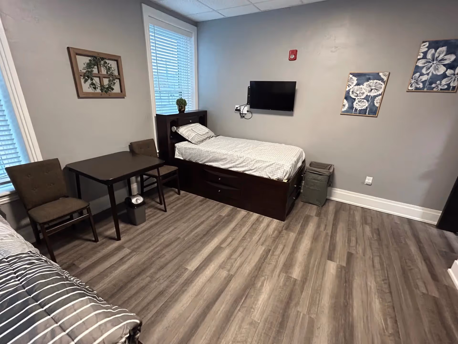 A clean and simple bedroom with two single beds, a small dark wooden table with two chairs, a wall-mounted TV, and decorative wall art including a framed cross with a wreath and floral prints. The room has wood-look flooring and light gray walls with two windows covered by white blinds.