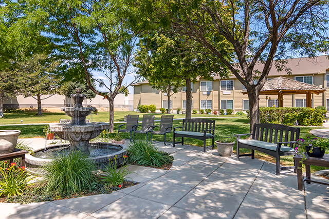 Outdoor seating area in a garden with benches and chairs arranged around a multi-tiered stone fountain. Trees provide shade and a building with multiple windows is visible in the background under a clear blue sky.