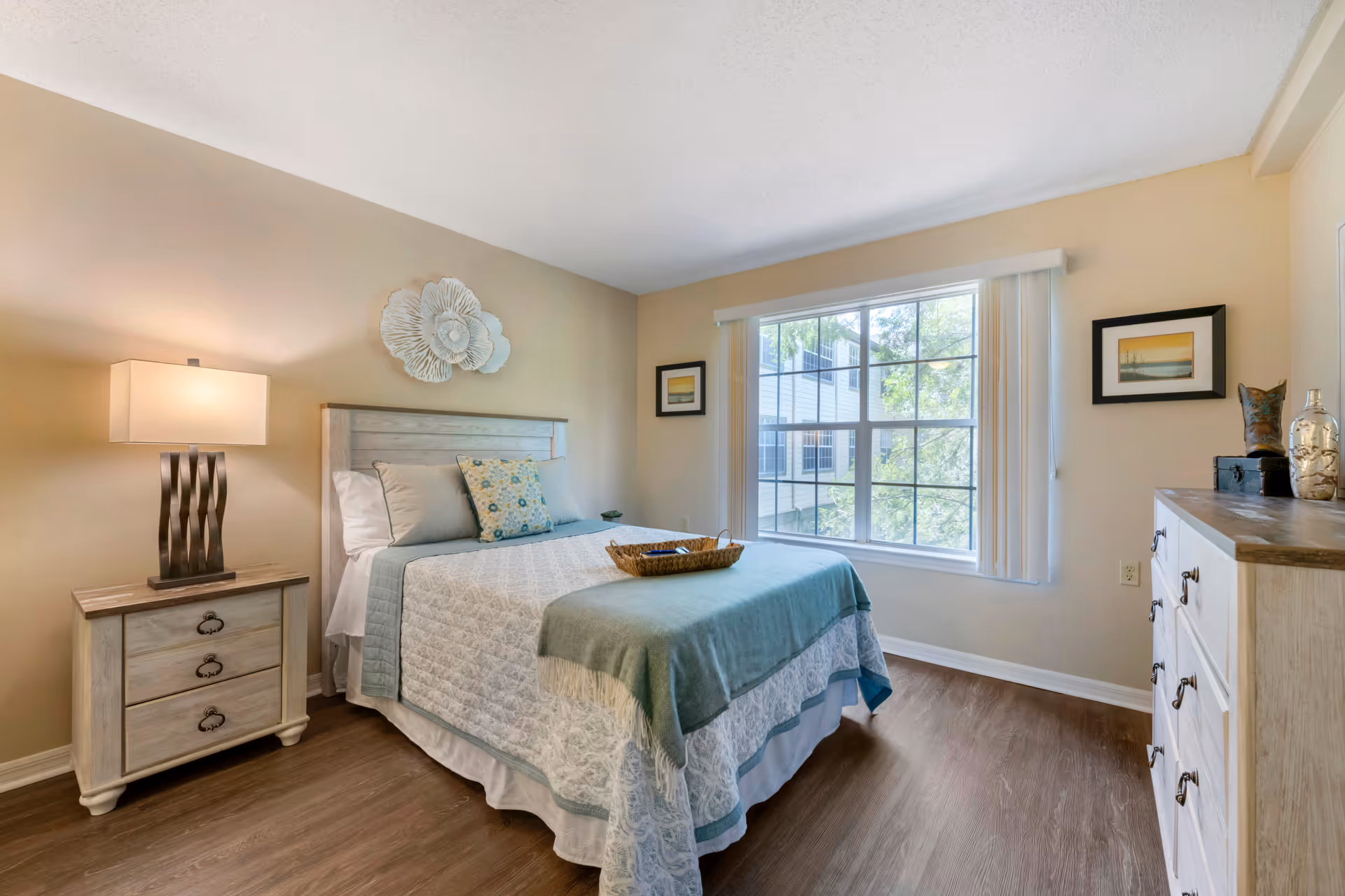 A bright and cozy bedroom with a large window letting in natural light. The room features a bed with a light blue and white quilt, decorative pillows, a wooden nightstand with a modern lamp, and a wooden dresser with decorative items on top. The walls are painted beige and decorated with framed pictures and a floral wall art piece above the bed.