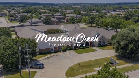 Aerial view of Meadow Creek Senior Living facility showing a large building surrounded by trees, parking areas, and a residential neighborhood in the background under a clear sky.