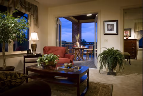 Cozy living room with a red armchair, wooden coffee table with decorative items, a lamp on a side table, and plants. The room opens to a balcony with a table and chairs, overlooking an outdoor view at dusk. A doorway leads to another room with a dresser and framed artwork on the walls.