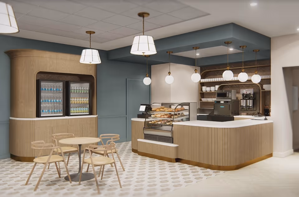 A modern cafe area inside a senior living facility featuring a display case with pastries, a counter with a cash register, shelves with cups and plates, a coffee machine, a refrigerator stocked with bottled drinks, and a small round table with four wooden chairs. The space is decorated with pendant lights and has a patterned tile floor.