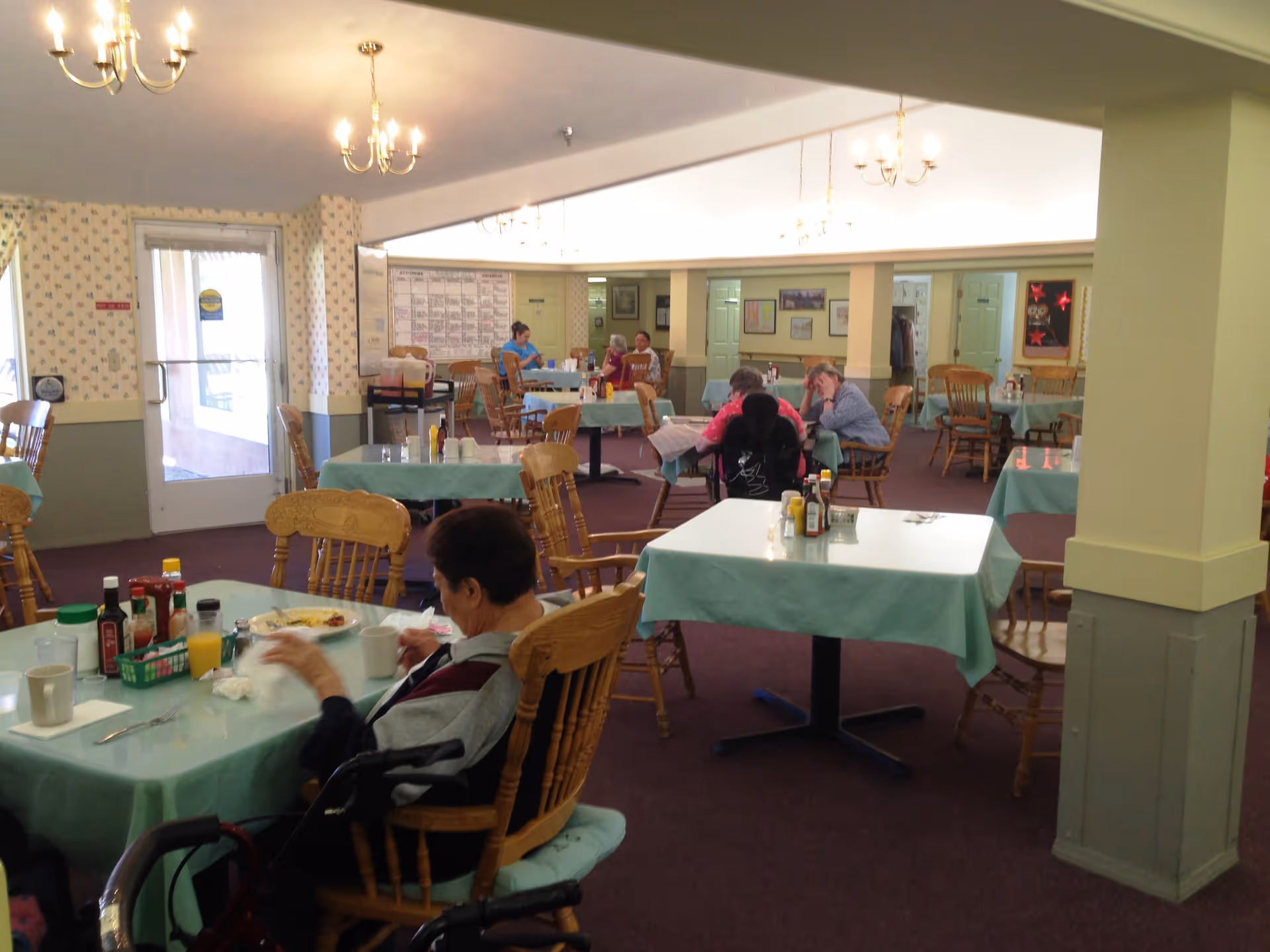 A dining room in an assisted living facility with several elderly residents seated at tables covered with light blue tablecloths. The room has wooden chairs, chandeliers hanging from the ceiling, and a door leading outside. Condiments and drinks are on the tables, and an activity calendar is visible on the far wall.