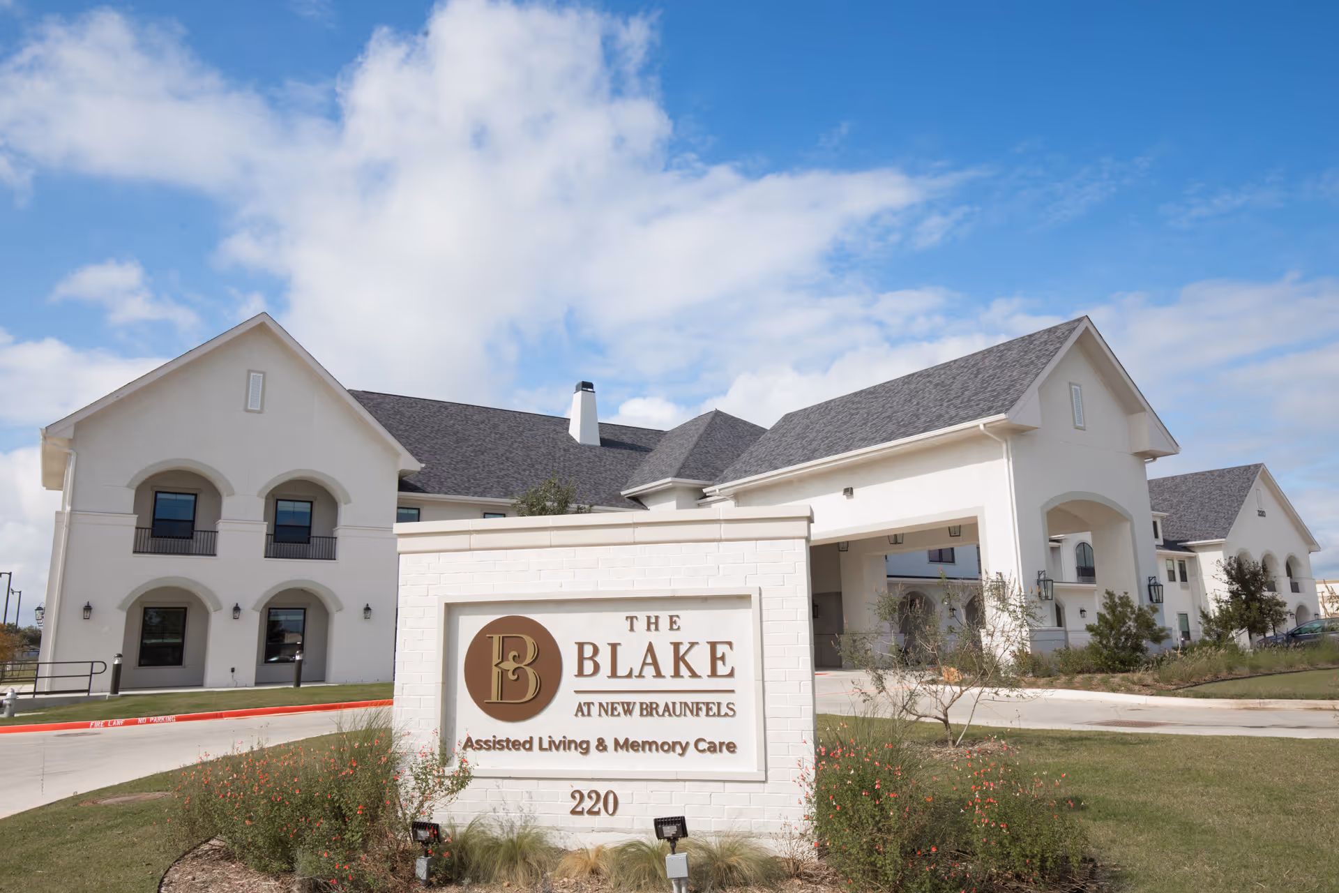 Exterior view of The Blake at New Braunfels assisted living and memory care facility with a large white sign in front displaying the facility name and address, surrounded by landscaping and a clear blue sky.