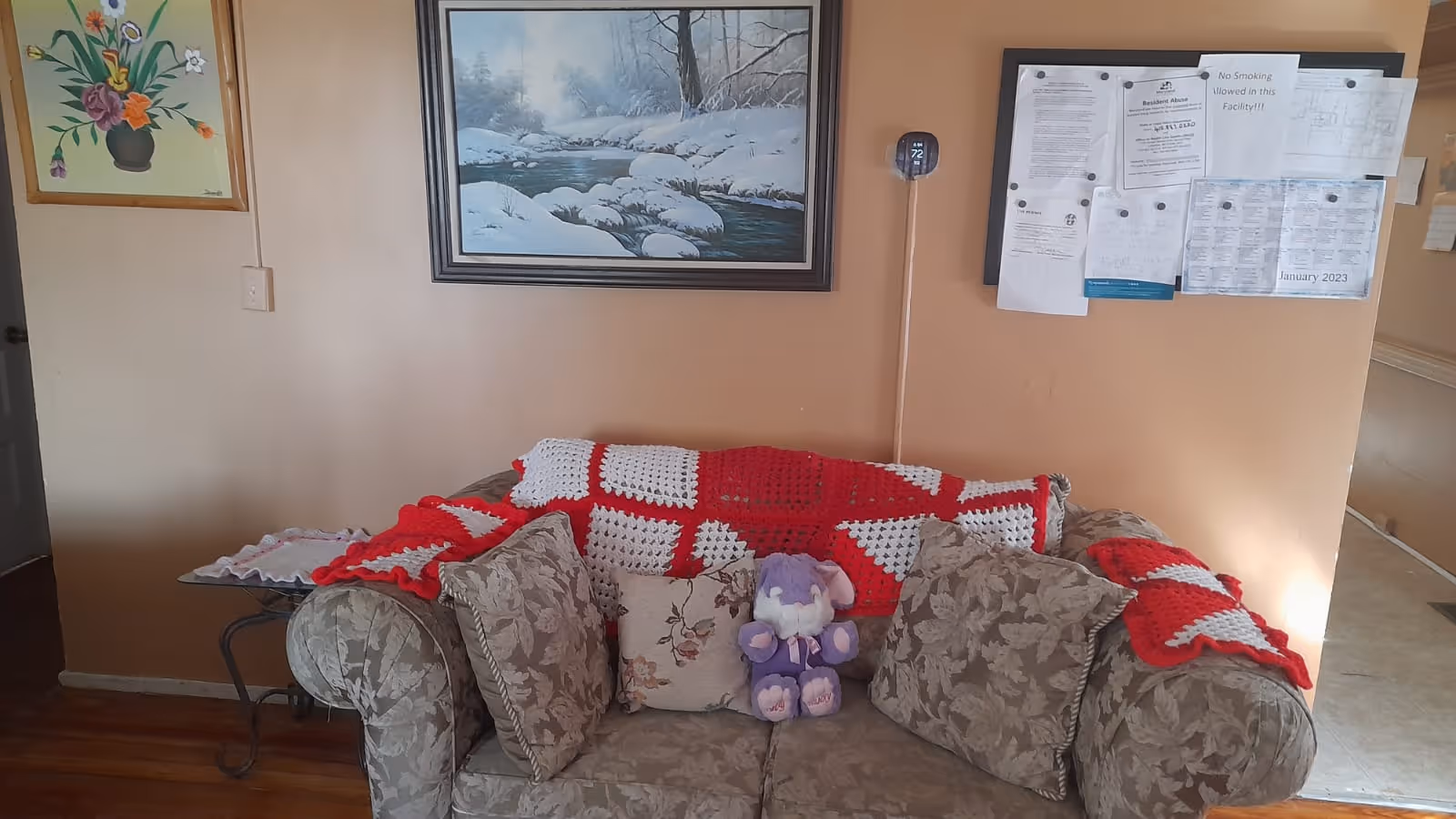 A floral-patterned sofa with a red-and-white crocheted throw, decorative pillows and a purple stuffed elephant sits against a wall with framed artwork and a bulletin board.