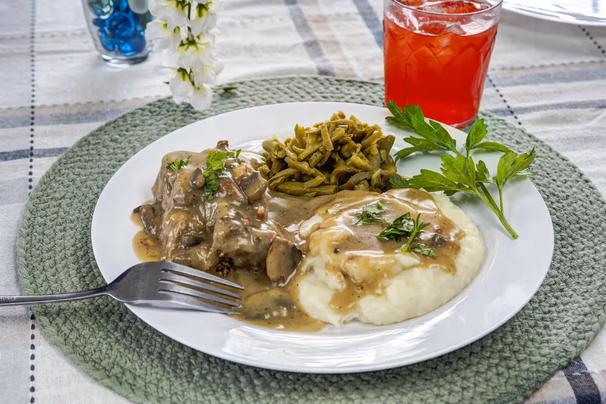 A white plate with a serving of mashed potatoes topped with brown gravy and chopped parsley, green beans, and a portion of meat covered in mushroom gravy. A fork rests on the plate. In the background, there is a glass of red beverage with ice on a green woven placemat on a checkered tablecloth.