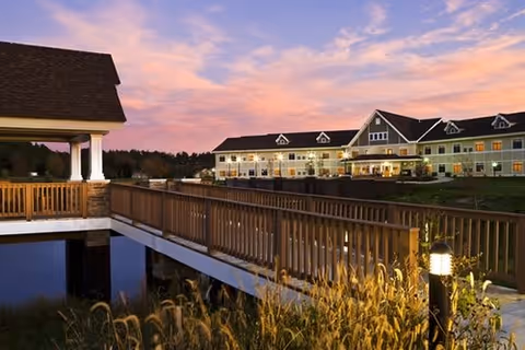 A senior living facility building seen at sunset with an illuminated wooden bridge over a pond and surrounding landscaping.