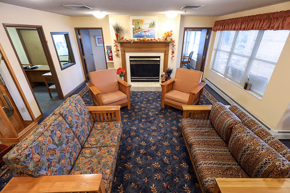 A cozy living room with two patterned sofas and two cushioned armchairs arranged around a fireplace. The fireplace mantel is decorated with autumn-themed ornaments and a painting above it. There is a large window with blinds and a valance on the right side, and doorways leading to other rooms on the left and center background.