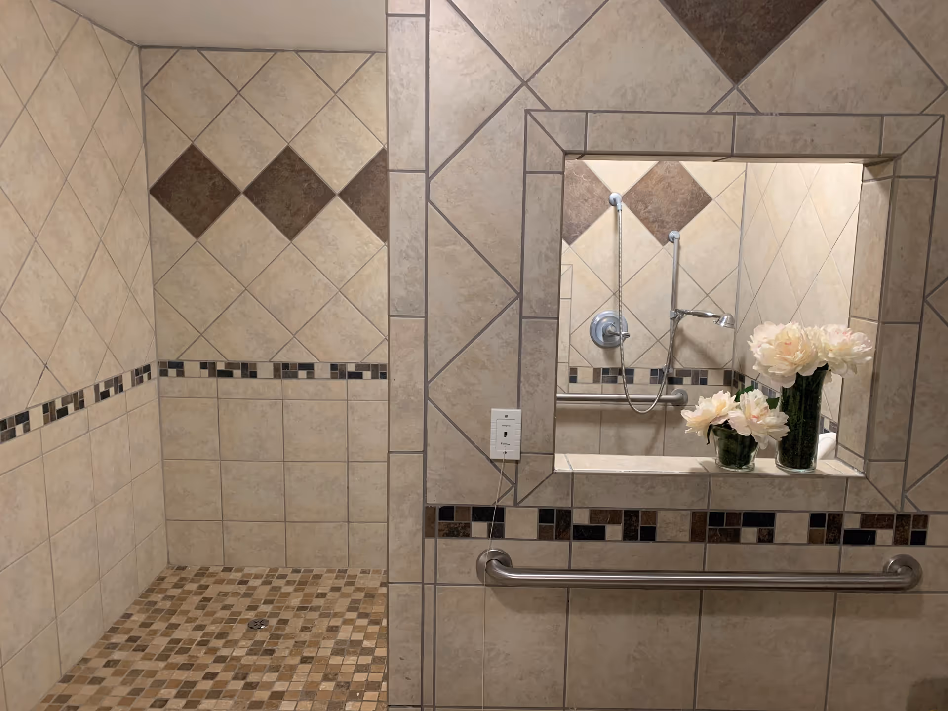 Tiled accessible shower area with grab bars and a recessed ledge/mirror holding vases of flowers and decorative tile accents.