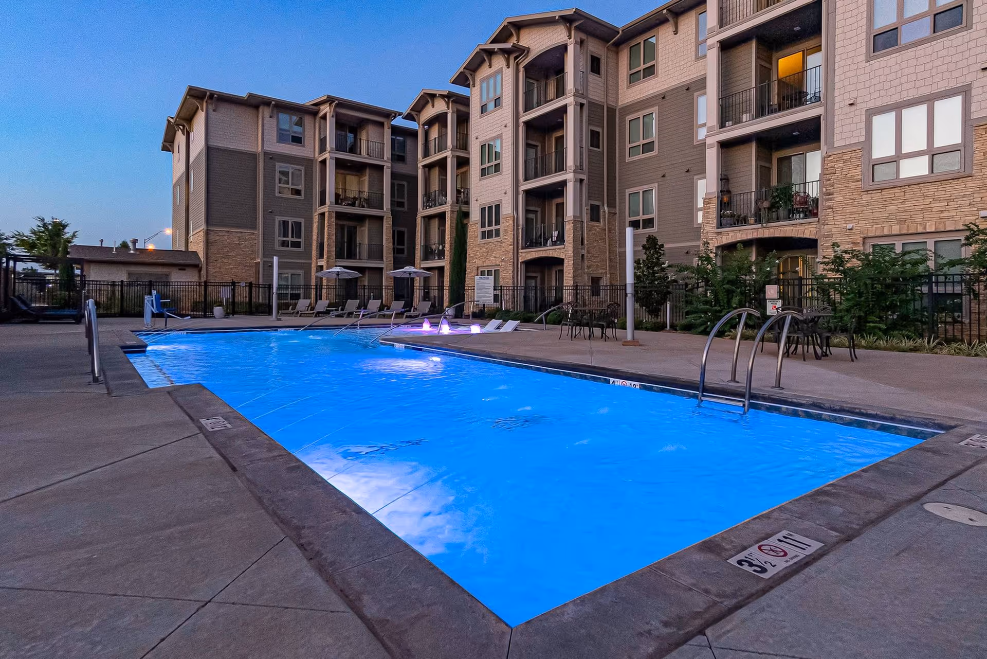 Outdoor swimming pool with blue water and small fountains, surrounded by a concrete deck with lounge chairs and tables. In the background, there is a multi-story residential building with balconies and windows during dusk.