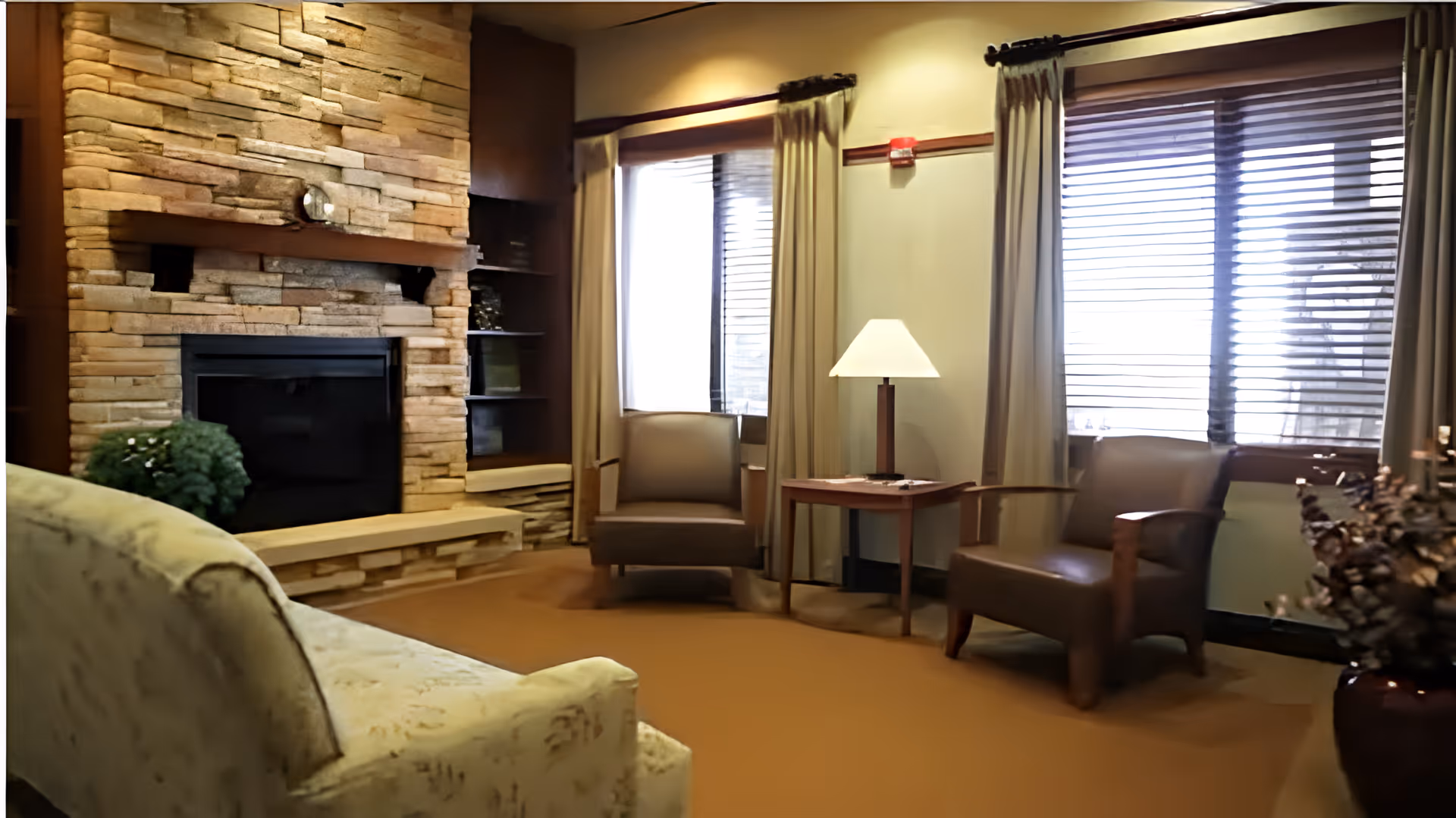 A cozy living room area with a stone fireplace, two armchairs, a side table with a lamp, and a sofa. The room has large windows with blinds and curtains, and a potted plant is visible near the fireplace.