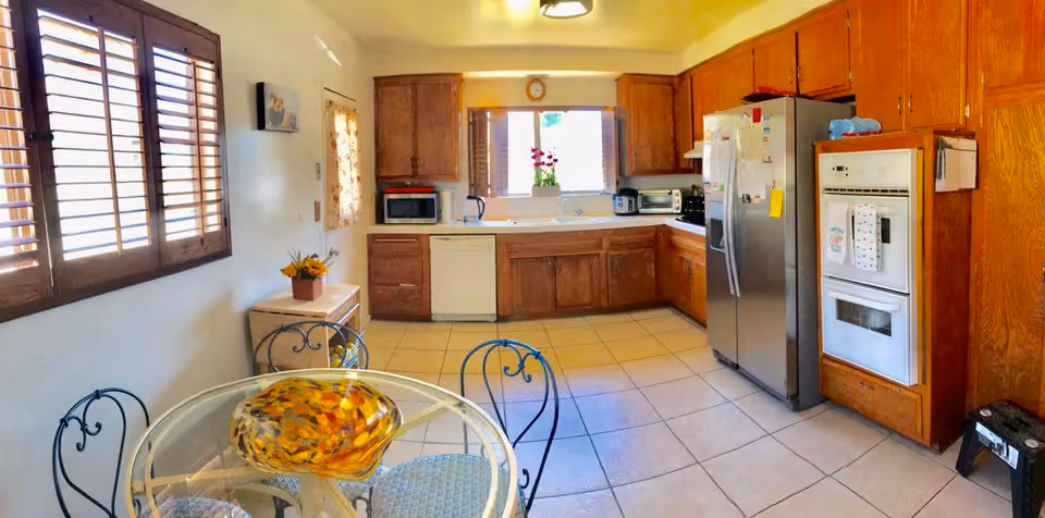 A bright kitchen with wooden cabinets, a stainless steel refrigerator, built-in oven, microwave, dishwasher, and various small appliances on the countertops. There is a round glass dining table with four metal chairs in the foreground, and windows with wooden shutters letting in natural light.