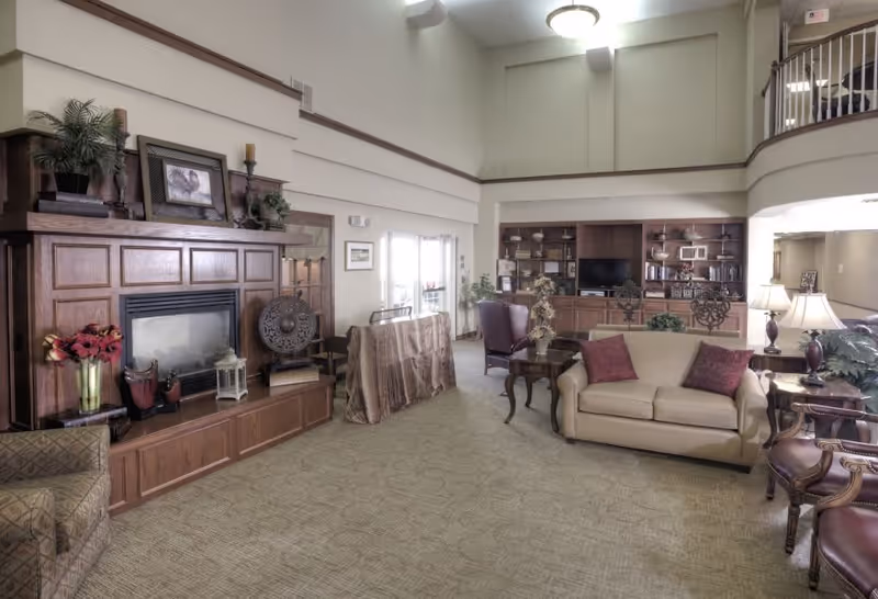 A spacious and well-lit living room area in a senior living facility with a beige sofa, armchairs, wooden coffee tables, and a large wooden built-in shelving unit with a television. There is a fireplace with decorative items on the mantel, including plants, candles, and framed pictures. The room has high ceilings and carpeted floors, with large windows allowing natural light to enter.