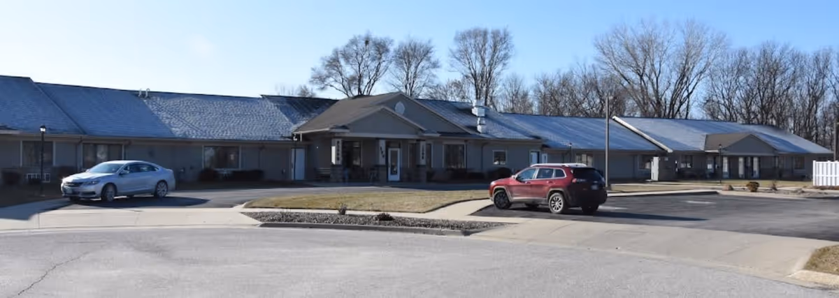 Exterior view of a single-story assisted living facility building with a pitched roof, several windows, and a covered entrance. Two cars are parked in front on a paved driveway, with leafless trees in the background under a clear sky.