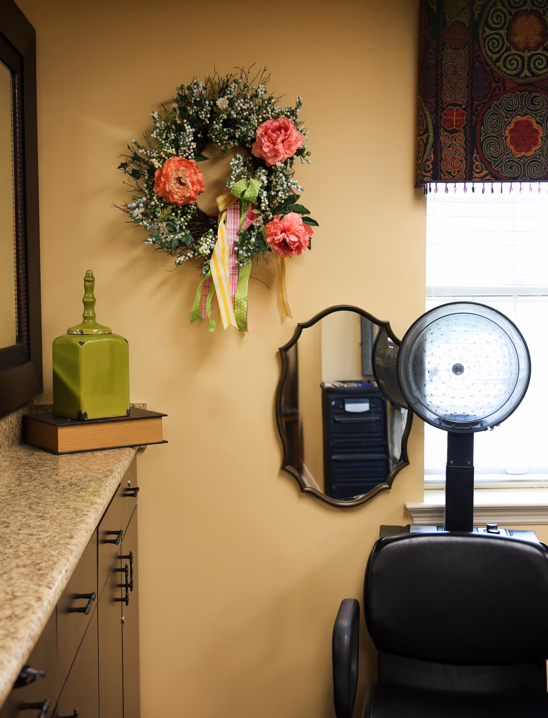 A salon corner with a hooded hair dryer and chair beneath a mirror, a decorative wreath on a beige wall, and countertop decor.