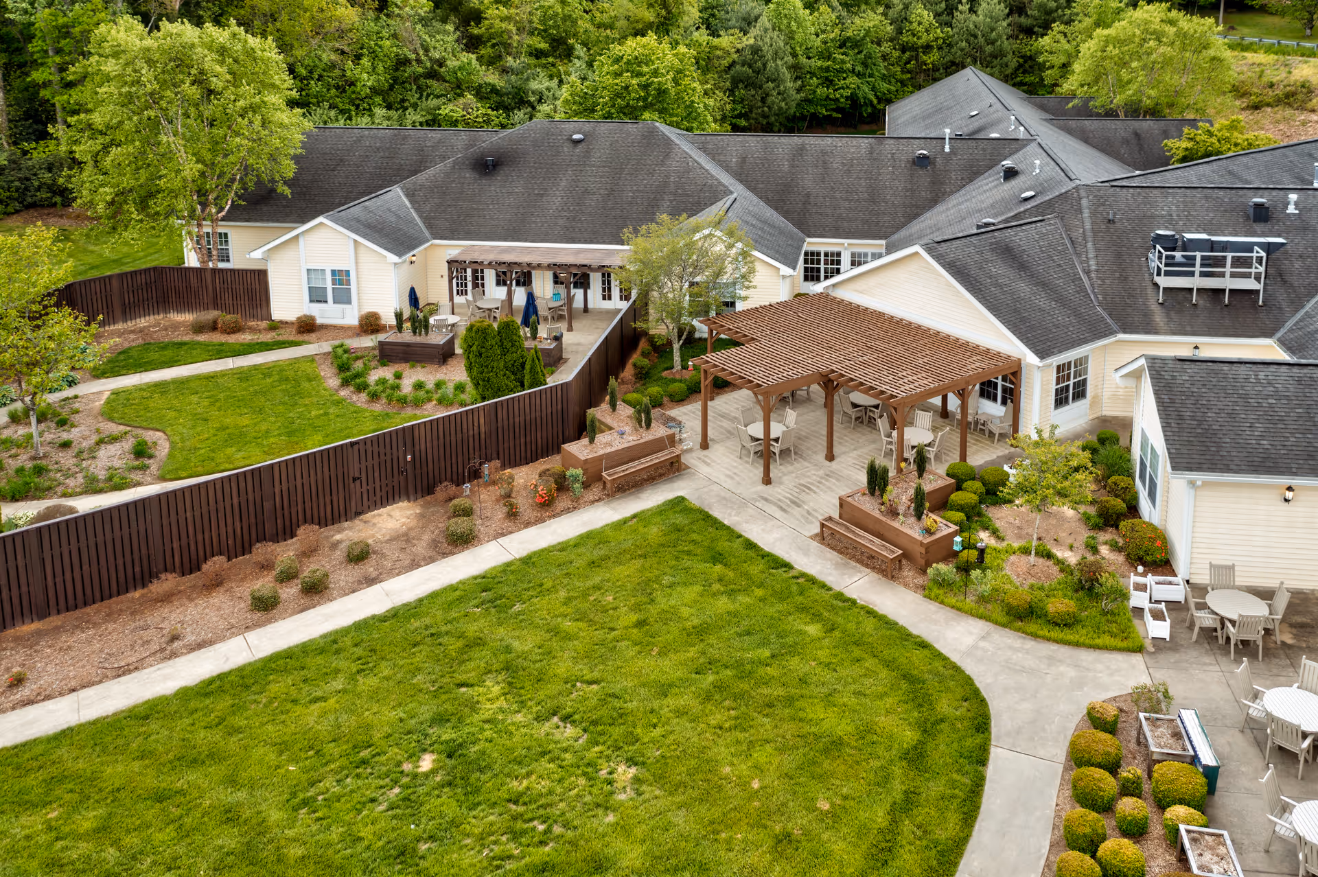 Aerial view of an assisted living facility showing a large green lawn, landscaped garden areas, and multiple outdoor seating areas with pergolas and tables. The building has a gray roof and light-colored exterior walls, surrounded by trees and greenery.