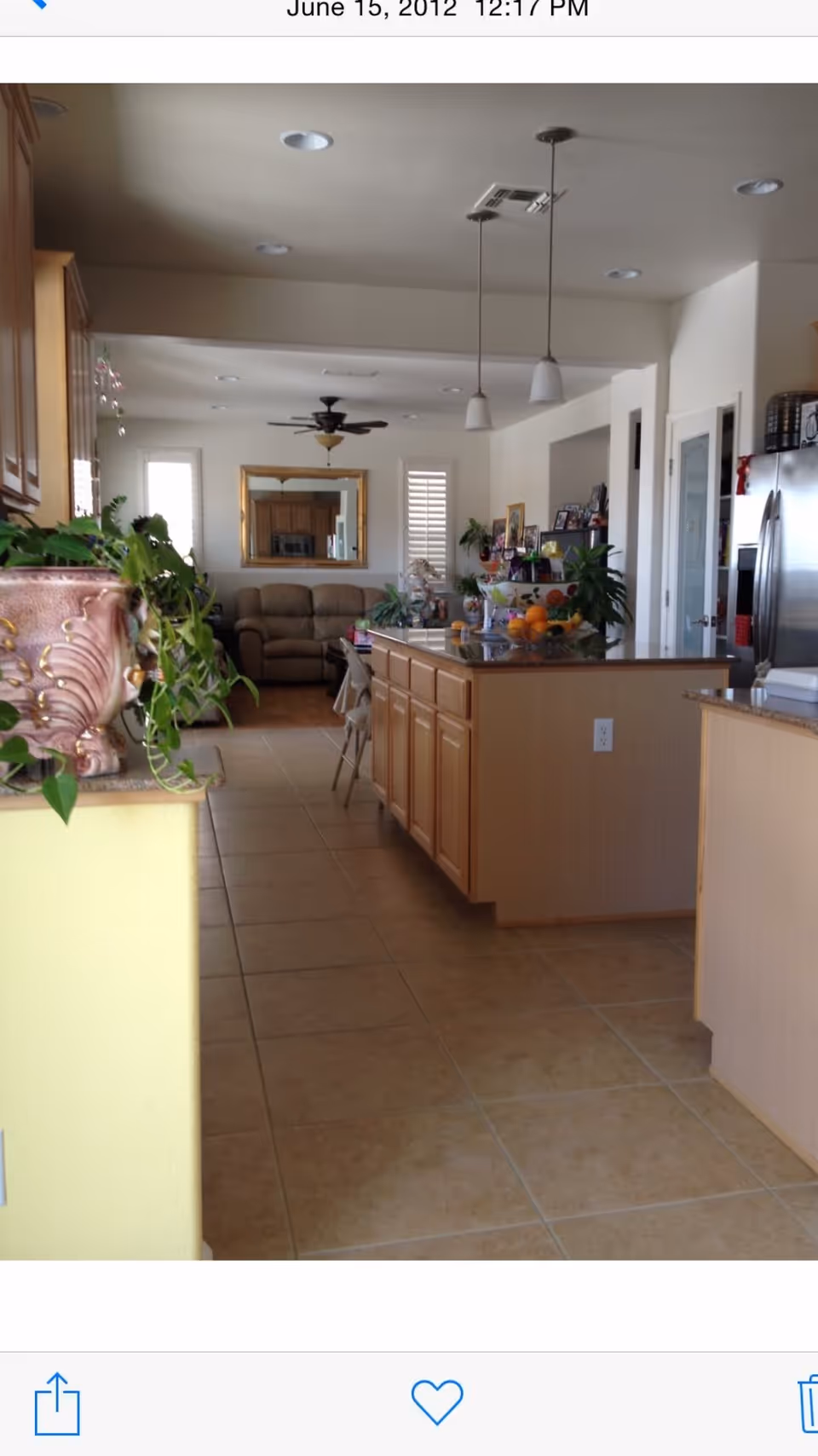 Interior view of a home showing a kitchen area with a large island countertop, wooden cabinets, hanging pendant lights, and a stainless steel refrigerator. In the background, there is a living room with a beige sofa, a ceiling fan, a large mirror on the wall, and windows with shutters. There are several plants and decorative items on the kitchen island and counters.