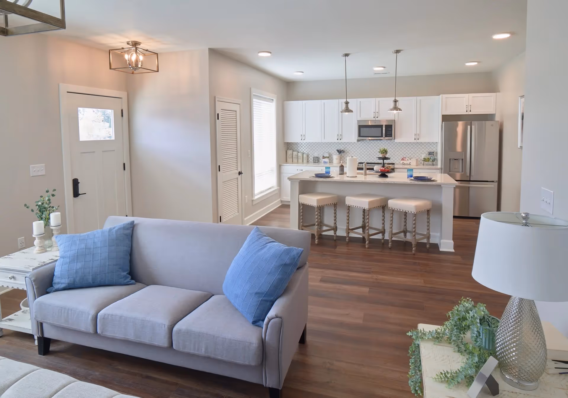 Open-plan living room with a gray sofa and blue pillows facing a kitchen island with three stools in a bright modern interior.