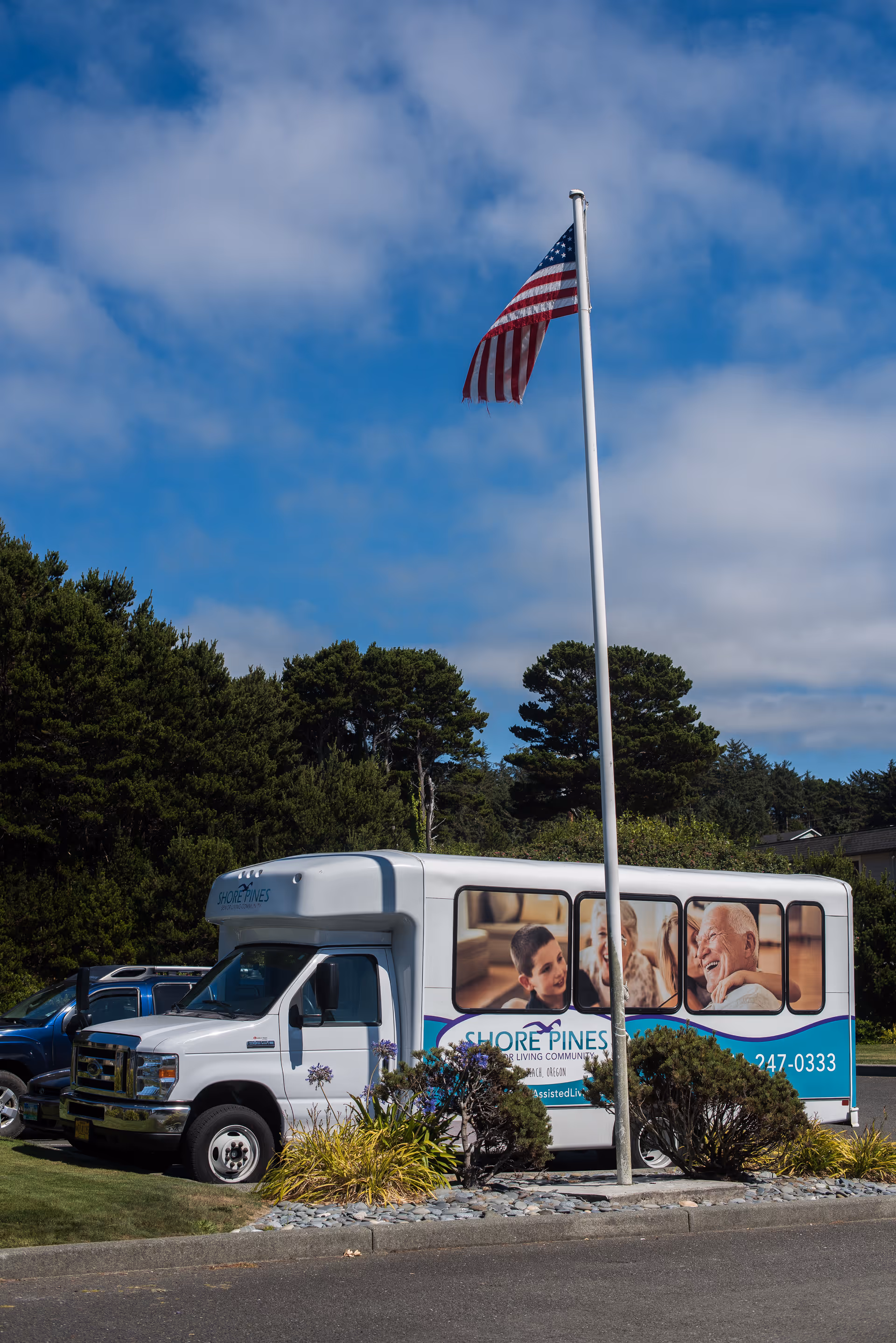 A white Shore Pines Senior Living Community shuttle bus parked near a flagpole with an American flag. The bus features images of smiling elderly people and caregivers on its side. The background includes green trees and a partly cloudy blue sky.