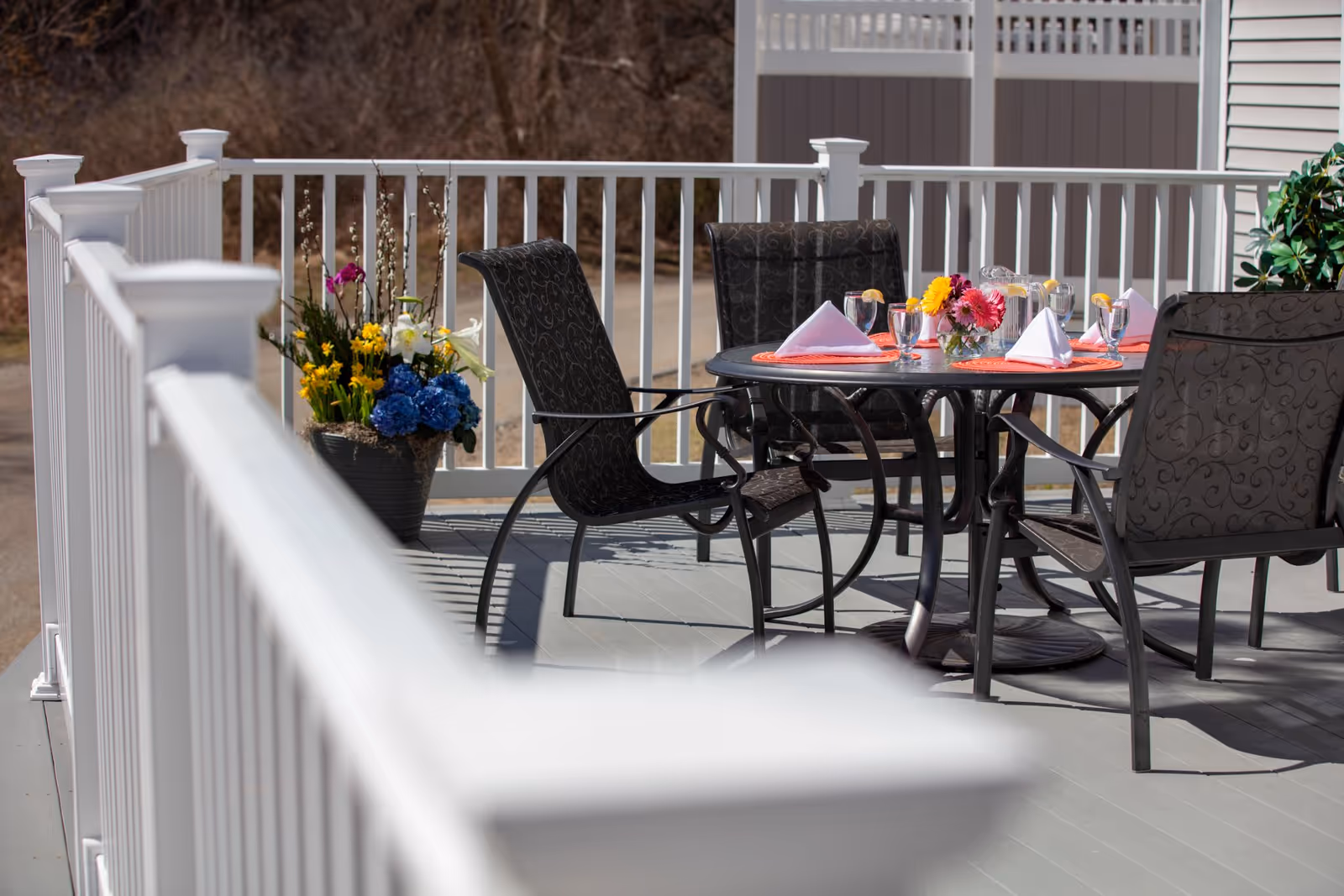 Outdoor patio area with a round table set for four, featuring black cushioned chairs, orange placemats, folded white napkins, glasses of water with lemon slices, a pitcher, and a small vase of colorful flowers. The patio is enclosed by a white railing and has a large flower pot with various flowers in the corner.