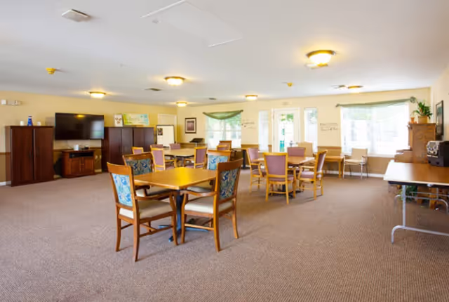 A spacious senior living common area with multiple wooden tables and chairs arranged on a carpeted floor. The room has beige walls, several windows letting in natural light, a large flat-screen TV mounted on a cabinet, and various pieces of furniture including a sideboard and a folding table. The ceiling has recessed lighting fixtures.