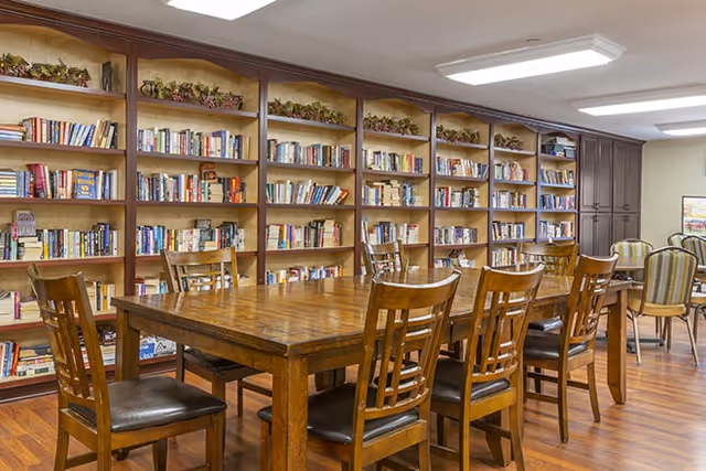 A well-lit room with wooden bookshelves filled with books along the wall, a large wooden table surrounded by wooden chairs with dark cushions, and additional seating with striped upholstered chairs in the background.