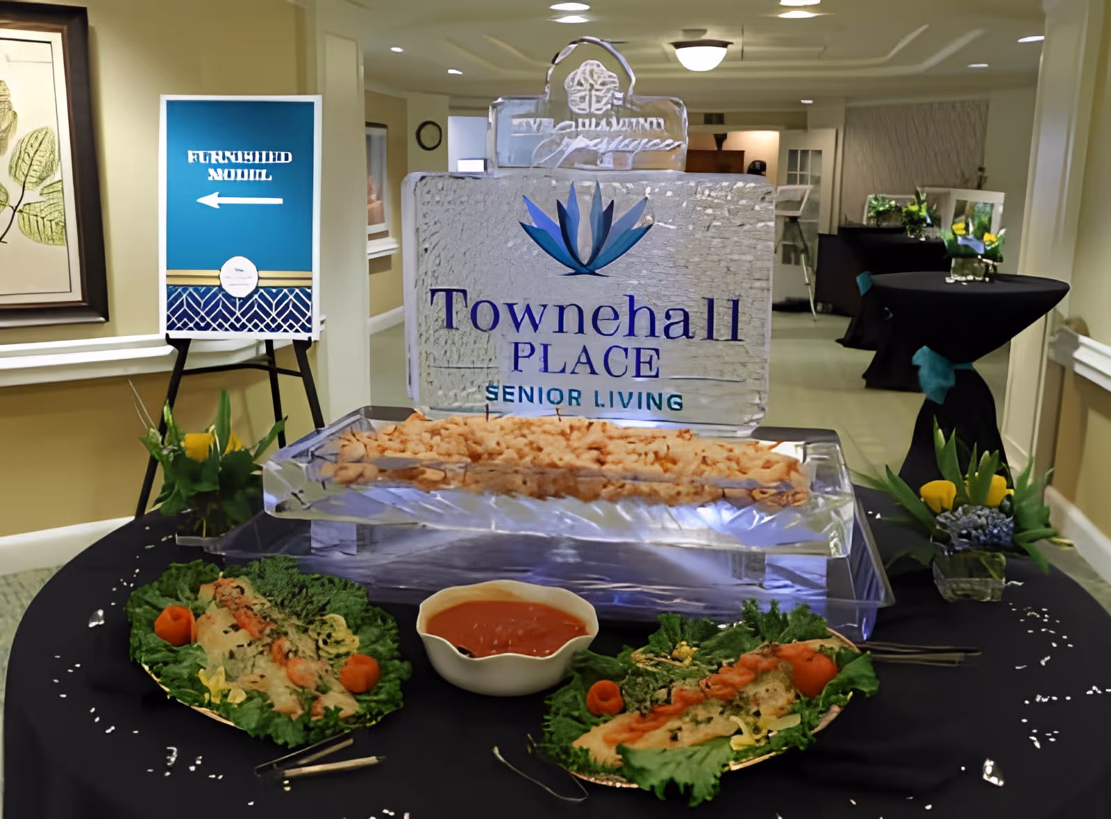 Buffet table in a facility lobby with an ice sculpture sign reading 'Townehall Place Senior Living' and plated appetizers.