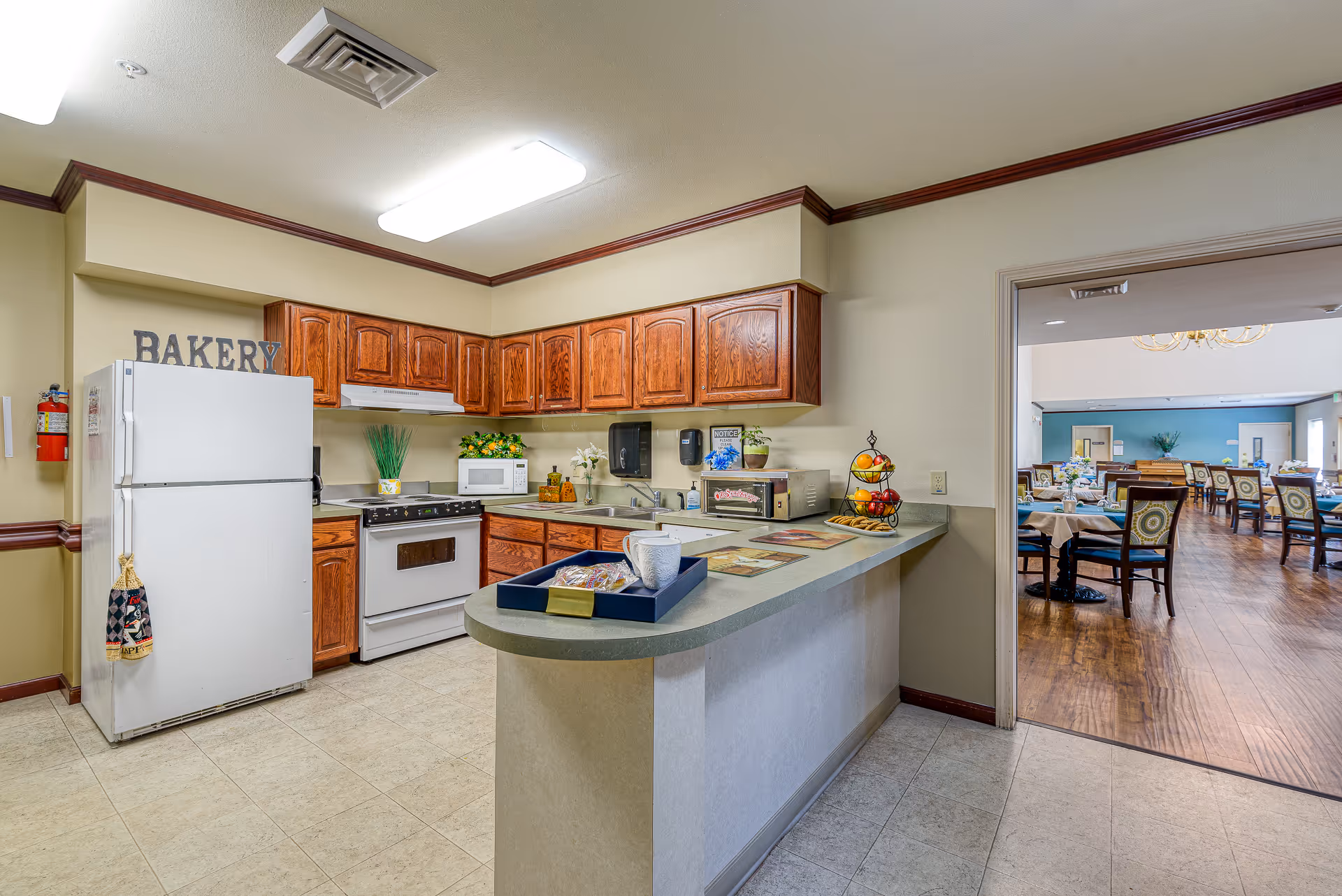 A kitchen area with wooden cabinets, a white refrigerator, stove, microwave, and a countertop with a tray holding a cup and snacks. The word 'BAKERY' is displayed above the refrigerator. Through an open doorway, a dining room with tables and chairs is visible.