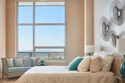 Bright bedroom with a large window overlooking water, a bed with patterned bedding and teal pillows, a bench by the window and decorative wall art.