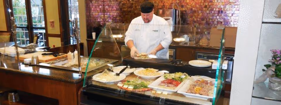 A chef prepares food behind a glass-covered buffet counter with trays of salads and cold dishes in a dining area.