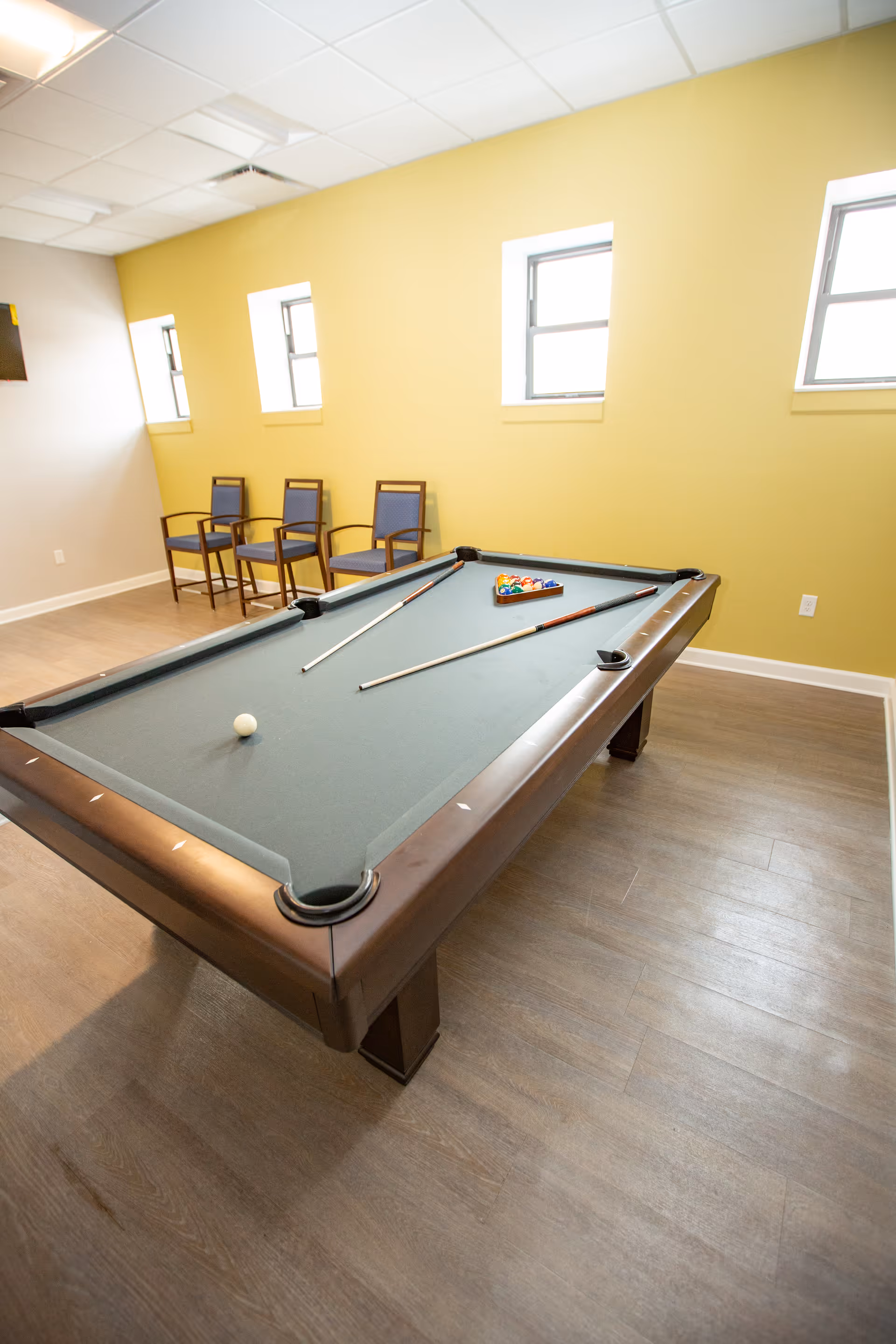 A room with a pool table set up for a game, featuring two pool cues and a rack of billiard balls. The room has three chairs lined up against a yellow wall with three windows, and the floor is covered with wood-like flooring.
