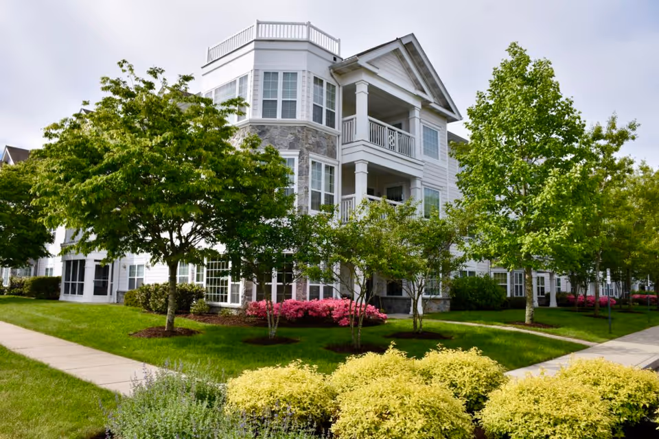 Exterior view of a multi-story senior living facility building with white siding and stone accents, surrounded by well-maintained green lawns, trees, and colorful flowering shrubs under a cloudy sky.
