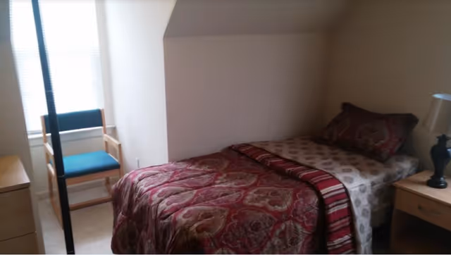 Small bedroom with a bed, quilt, nightstand, lamp, and a chair by the window.